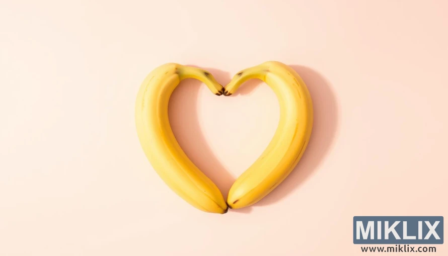 Heart-shaped arrangement of ripe yellow bananas against a soft, pastel-colored background. The bananas are slightly angled to form a symmetrical heart shape, with a warm, natural lighting casting gentle shadows. The overall composition conveys a sense of health, wellness, and the cardiovascular benefits of eating bananas. The image has a clean, minimalist aesthetic with a focus on the vibrant fruit and its symbolic representation of heart health.